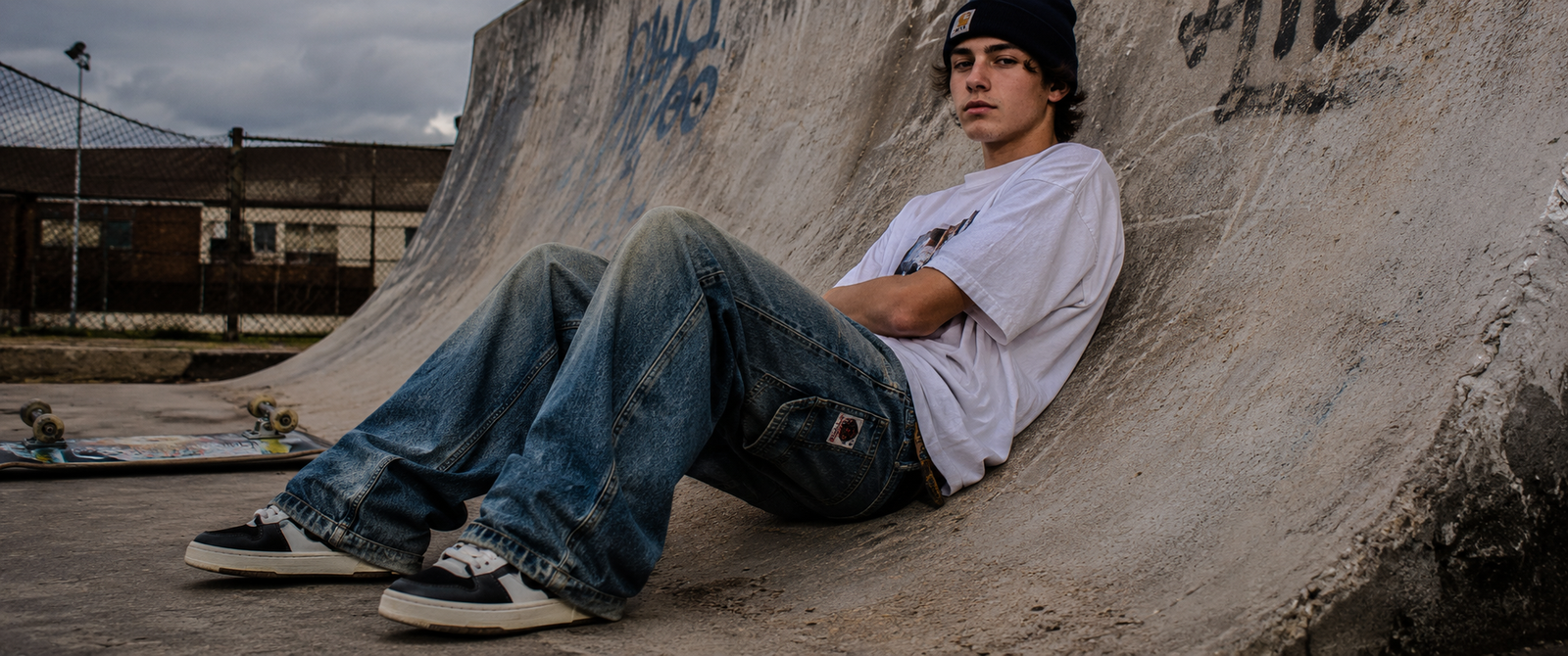 Youth skater leaning against a concrete halfpipe in a skatepark, wearing vintage faded indigo baggy jeans and chunky skate sneakers, captured in a moody 1990s-inspired street portrait with gritty urban lighting.